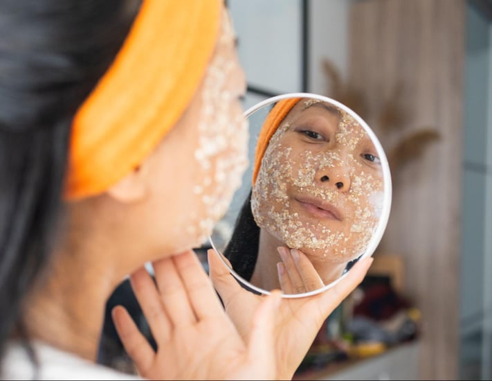 woman exfoliating skin while looking in the mirror